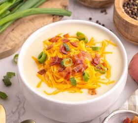loaded potato soup, A bowl of creamy potato soup topped with shredded cheddar cheese chopped bacon and sliced green onions Fresh potatoes green onions and bowls of spices are arranged around it on a white marble surface