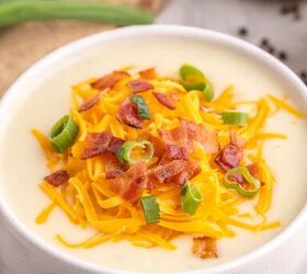loaded potato soup, A white bowl filled with creamy soup topped with shredded cheddar cheese crispy bacon pieces and sliced green onions Green onions and a wooden bowl are visible in the background