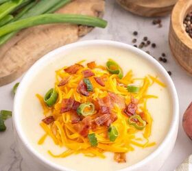 loaded potato soup, A bowl of creamy potato soup topped with shredded cheddar cheese crispy bacon bits and sliced green onions Sliced green onions a bowl of salt and a cutting board are in the background