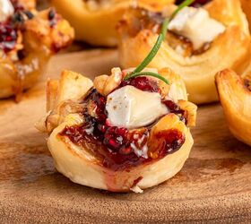 cranberry brie puff pastry bites gluten free, A close up of a baked gluten free puff pastry cup filled with melted brie cheese cranberry jam walnuts and garnished with a sprig of rosemary sitting on a wooden surface