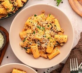 creamy italian sausage pasta with spinach, A bowl of rigatoni creamy pasta topped with ground meat fresh herbs and grated cheese placed on a light table with bowls of pasta grated cheese and fresh parsley nearby