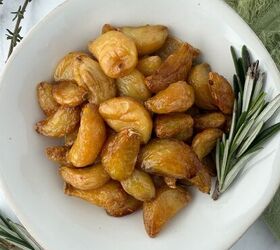 garlic confit with thyme and rosemary, A white plate filled with golden brown garlic confit cloves garnished with fresh rosemary sprigs on the side placed on a light surface with a green cloth nearby
