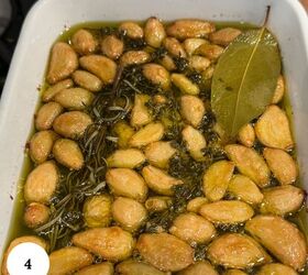 garlic confit with thyme and rosemary, A white dish filled with garlic confit roasted garlic cloves submerged in olive oil garnished with fresh rosemary and a bay leaf
