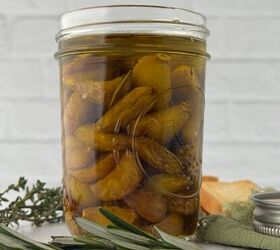 garlic confit with thyme and rosemary, A glass jar filled with yellow and green olives in oil and garlic confit is placed on a table surrounded by sprigs of fresh herbs bread and a green napkin against a white brick background