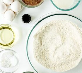 chocolate caramel cake recipe rich moist easy, Overhead view of chocolate cake ingredients arranged on a white surface including a large bowl of flour cocoa powder milk in a measuring cup eggs oil vanilla extract sugar and baking powder in small glass bowls