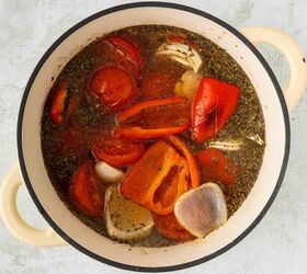 roasted red pepper and tomato soup, A cream colored pot filled with tomato halves red bell pepper pieces garlic cloves herbs and broth all simmering together on a light gray background