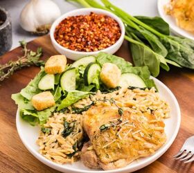 creamy chicken orzo bake one pan dinner, A plate of chicken orzo bake with crispy roasted chicken served atop creamy orzo pasta with spinach accompanied by a green salad with cucumber slices and croutons Bowls of red pepper flakes and fresh herbs are nearby