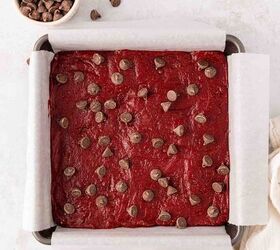 easy red velvet cake mix brownies, A square baking pan lined with parchment paper is filled with unbaked red velvet brownie batter topped with chocolate chips A small bowl of chocolate chips sits nearby on a white surface