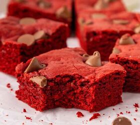 easy red velvet cake mix brownies, Red velvet brownies topped with chocolate chips are arranged on parchment paper with one brownie in the foreground showing a large bite taken out of it and scattered crumbs around