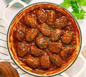greek beef meatballs in tomato sauce soutzoukakia, Round white pot filled with Greek Beef Meatballs or Soutzoukakia in rich red tomato sauce garnished with chopped parsley A striped towel wooden spoon and fresh parsley leaves are nearby on a light colored tabletop
