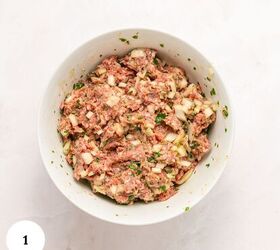 greek beef meatballs in tomato sauce soutzoukakia, A white bowl filled with a mixture for Greek Beef Meatballs combining ground meat chopped onions herbs and seasonings sits on a light colored surface A white circle with the number 1 is in the lower left corner