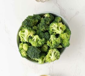 air fryer broccoli, A bowl of fresh broccoli florets a small dish of olive oil and a scalloped dish containing salt pepper and granulated seasonings are arranged on a white marble surface