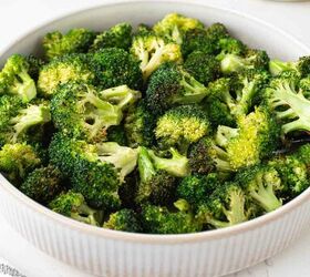 air fryer broccoli, A bowl filled with roasted broccoli florets sits on a white marble surface with a stack of plates and forks in the background and a striped cloth nearby