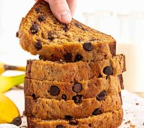 chocolate chip banana bread ultra moist easy, A hand lifts a slice from a stack of thick chocolate chip filled banana bread slices on parchment paper Bananas and glass bottles of milk are visible in the background