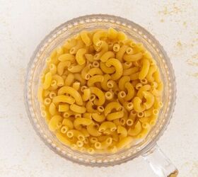 microwave mac and cheese, A clear glass measuring cup filled with uncooked elbow macaroni noodles viewed from above on a light textured surface