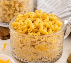 microwave mac and cheese, A clear glass mug filled with creamy macaroni and cheese sits on a table next to a striped cloth napkin and a fork with a bowl of shredded cheese partially visible nearby