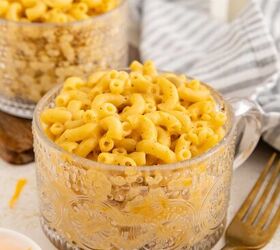 microwave mac and cheese, A clear glass mug filled with creamy macaroni and cheese sits on a table next to a gold fork a striped gray napkin and another mug of macaroni in the background Shredded cheese is in a small bowl nearby