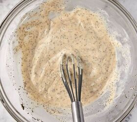 creamy coleslaw with celery seed, A glass bowl filled with a creamy mixture containing visible black specks of celery seed A metal whisk rests in the bowl The background is a white marble surface and a dark cloth is partially visible in the top left corner