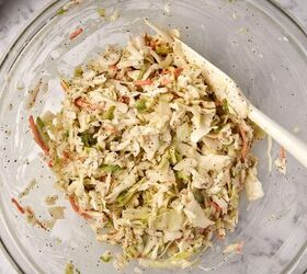 creamy coleslaw with celery seed, A clear glass bowl filled with coleslaw featuring shredded cabbage carrots and a sprinkle of black pepper A white spatula rests inside the bowl The background shows a marble surface and a gray cloth in the corner