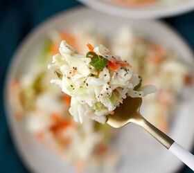 creamy coleslaw with celery seed, Close up of a fork holding a bite of coleslaw with cabbage carrots and green onions The dish is on a blurred plate in the background set on a teal cloth The coleslaw appears creamy and speckled with black pepper