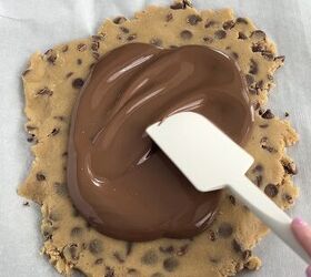 easy no bake cookie dough bark edible safe to eat, A hand spreads melted chocolate over a flattened round of chocolate chip cookie dough using a white spatula all on parchment paper atop a gray surface