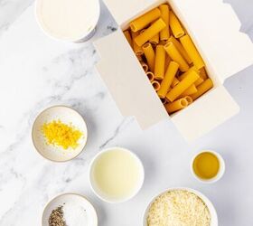 lemon ricotta pasta without heavy cream, A flat lay of dry rigatoni pasta in an open box grated cheese ricotta olive oil lemon zest black pepper and salt in small bowls on a white marble surface