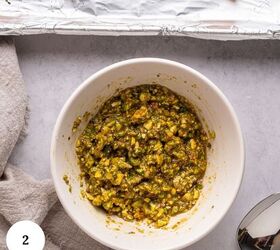 pistachio crusted salmon with lemon and za atar, A white bowl filled with a chunky green and yellow pesto mixture sits on a light countertop next to a silver spoon a beige cloth and a foil lined baking sheet with pistachio crusted salmon pieces A 2 label is in the bottom left corner