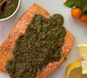pistachio crusted salmon with lemon and za atar, A cooked salmon pesto fillet topped with green sauce on a white plate garnished with lemon wedges yellow cherry tomatoes fresh basil and a small bowl of extra pesto with a wooden spoon