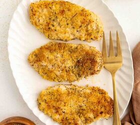 baked parmesan crusted chicken better than fried, Three breaded and baked chicken cutlets are arranged on a white oval plate with a gold fork beside them A small bowl of grated cheese and a bowl of salt are nearby on the table