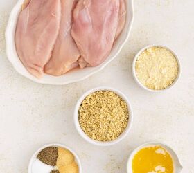 baked parmesan crusted chicken better than fried, Raw chicken breasts in a white dish surrounded by small bowls containing breadcrumbs grated Parmesan cheese melted butter and assorted spices on a light surface