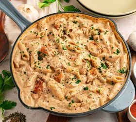 garlic parmesan pasta with chicken, A skillet filled with creamy garlic parmesan pasta with chicken pieces garnished with chopped parsley and black pepper Garlic herbs and empty plates are arranged around the dish on a light colored surface