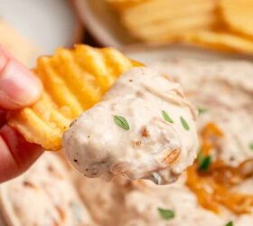 greek yogurt french onion dip with real caramelized onions, A hand holds a crinkle cut potato chip dipped in creamy Greek yogurt French onion dip garnished with small green herbs More chips and dip are visible in the blurred background