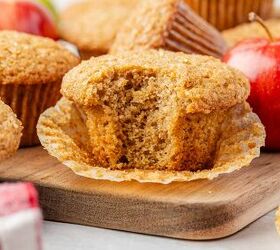 moist applesauce muffins, A close up of an applesauce muffin with a bite taken out resting on an unwrapped paper liner on a wooden board surrounded by whole muffins and a red apple