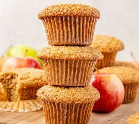moist applesauce muffins, Three apple muffins are stacked on a wooden board surrounded by red apples and more muffins with one muffin partially unwrapped and a red checked cloth visible in the corner