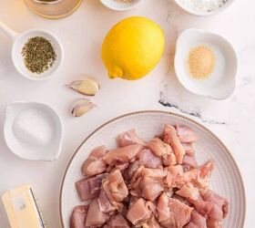 garlic butter chicken bites ready in 20 minutes, A plate of raw cubed chicken surrounded by a lemon garlic cloves butter a bottle of oil and small bowls of seasonings spices and flour on a white marble surface
